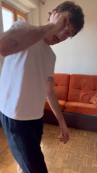 Uomo con maglietta bianca e pantaloncini scuri regola gli occhiali in una stanza luminosa con divano arancione e pavimento in parquet di legno.