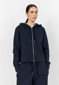 Jūras spēku krāsā zip-augšējā kapuce ar auklu kapuci, izgatavota no mīksta auduma. Izceļas ar teksturētu virsmu un ribotu piedurkņu apmalēm. Komplektā ar atbilstošām biksēm.