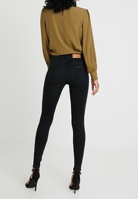 Jeans skinny negros de cintura alta, con un diseño de bolsillo trasero y un corte ajustado, combinados con una blusa marrón y tacones altos negros.