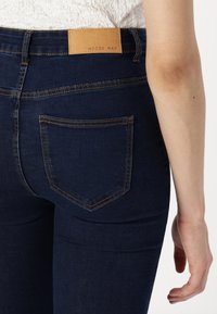 Dark blue denim jeans with a snug fit, featuring a leather patch labeled "NOISY MAY" and simple back pockets with contrasting stitching.