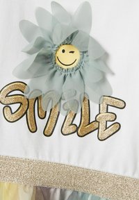 Tissu blanc arborant le mot "SOURIRE" en paillettes dorées. Une fleur bleu clair avec un visage souriant met en valeur le design, renforçant son attrait visuel.
