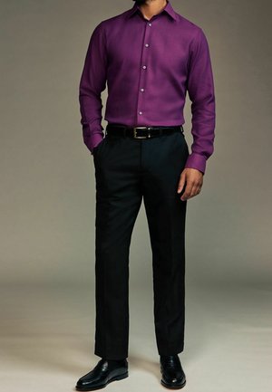 Man in een donkerpaars overhemd, zwarte nette broek, zwarte riem en zwarte nette schoenen, staand met één hand in zijn zak.