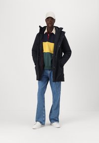 Parka nera con zip e coulisse, indossata sopra un maglione a righe multicolori, abbinata a jeans a gamba larga blu chiaro e sneakers bianche.