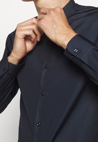 Marineblauw overhemd met knopen en een mandarin kraag, gemaakt van gladde stof en voorzien van glanzende zwarte knopen, deels dichtgeknoopt bij de hals.