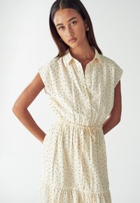 Světle béžové šaty s černými puntíky, krátké rukávy, zapínání na knoflíky v přední části a pružný pas. Design s volánkovou sukní. Měkká textilie.