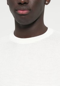 Λευκό πλεκτό πουλόβερ με ribbed υφή και στρογγυλή λαιμόκοψη. Κοντινή λήψη που εστιάζει στις λεπτομέρειες του υφάσματος. Ορατά μικρά σκουλαρίκια.