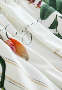 Tissu blanc à imprimé botanique avec des feuilles vertes, des fleurs rouges, et un texte cursif noir partiellement visible sur les plis.