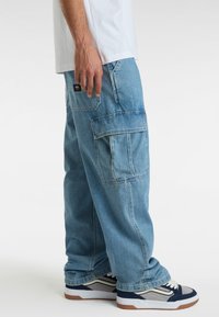 Jeans cargo blu con molte tasche, vestibilità comoda e dettagli cuciti. Abbinati a scarpe da ginnastica blu e grigie e a una maglietta bianca.