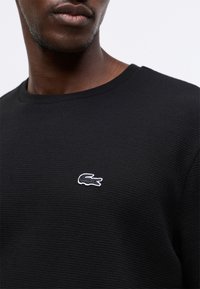 Close-up de uma pessoa a usar uma camisa preta de manga longa texturizada com um pequeno logótipo de crocodilo contornado a branco no peito.