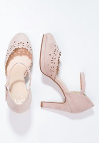 Para różowych zamszowych szpilek z paskami na kostce i wyciętymi kwiatowymi wzorami na białym tle.