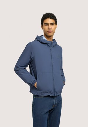 Ung mand iført en blå hættetrøje med lynlås og hænderne i lommerne samt mørkeblå jeans mod en ensfarvet baggrund.