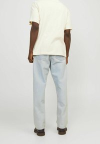 En person som bär en ljuskrämfärgad t-shirt med korta ärmar, ljusblå jeans och mörkbruna skor, står vänd bort från kameran mot en neutral bakgrund.