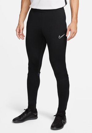 Homme portant des leggings de sport noirs Nike et des baskets noires Nike, debout devant un fond clair uni.