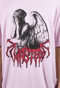 Růžové bavlněné tričko s grafikou anděla v černobílé barvě, která má opotřebovaný vzhled, a stylizovaným červeným nápisem "WASTED" ve spodní části.