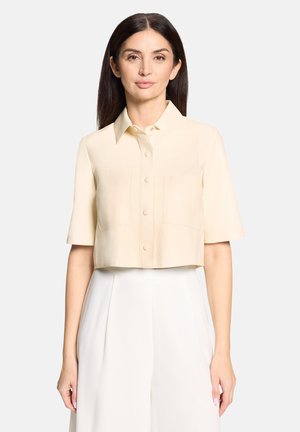 Frau mit langen dunklen Haaren, die ein beiges kurzärmeliges Hemd mit Knöpfen und weiße hoch taillierte Hosen trägt, steht vor einem schlichten Hintergrund.