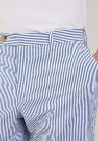 Pantalons rayés bleu et blanc avec une fermeture à bouton, présentant des poches latérales et un tissu léger et lisse. Coupe ajustée avec une ceinture propre.