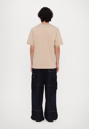 Persona con el cabello rizado y oscuro, vistiendo una camiseta beige de manga corta y pantalones cargo negros sueltos, de pie de espaldas sobre un fondo blanco liso.
