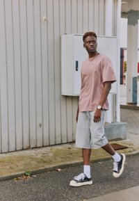Jeune homme portant un T-shirt rose oversize, un short en denim clair, des chaussettes blanches et des baskets noires, marchant près d'un mur en métal ondulé en plein air.