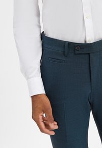 Dunkelgrüne, maßgeschneiderte Hose mit glatter Textur, Gürtelschlaufen und einer seitlichen Tasche. Kombiniert mit einem weißen Hemd mit Knöpfen.