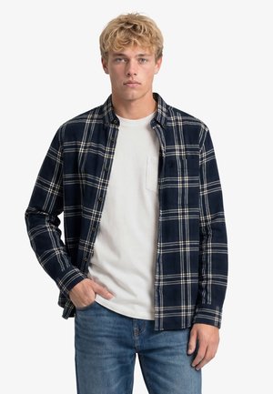 Jeune homme aux cheveux blonds porte une chemise à carreaux bleu marine par-dessus un t-shirt blanc et un jean bleu, debout avec une main dans la poche.