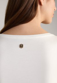 White short-sleeve top featuring a back circular hardware detail with a stylized logo. Smooth texture with a relaxed fit.