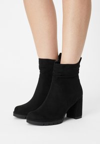 Bottines en suede noir avec un design plié, un talon épais haut et une semelle en caoutchouc. Texture douce et style minimaliste.