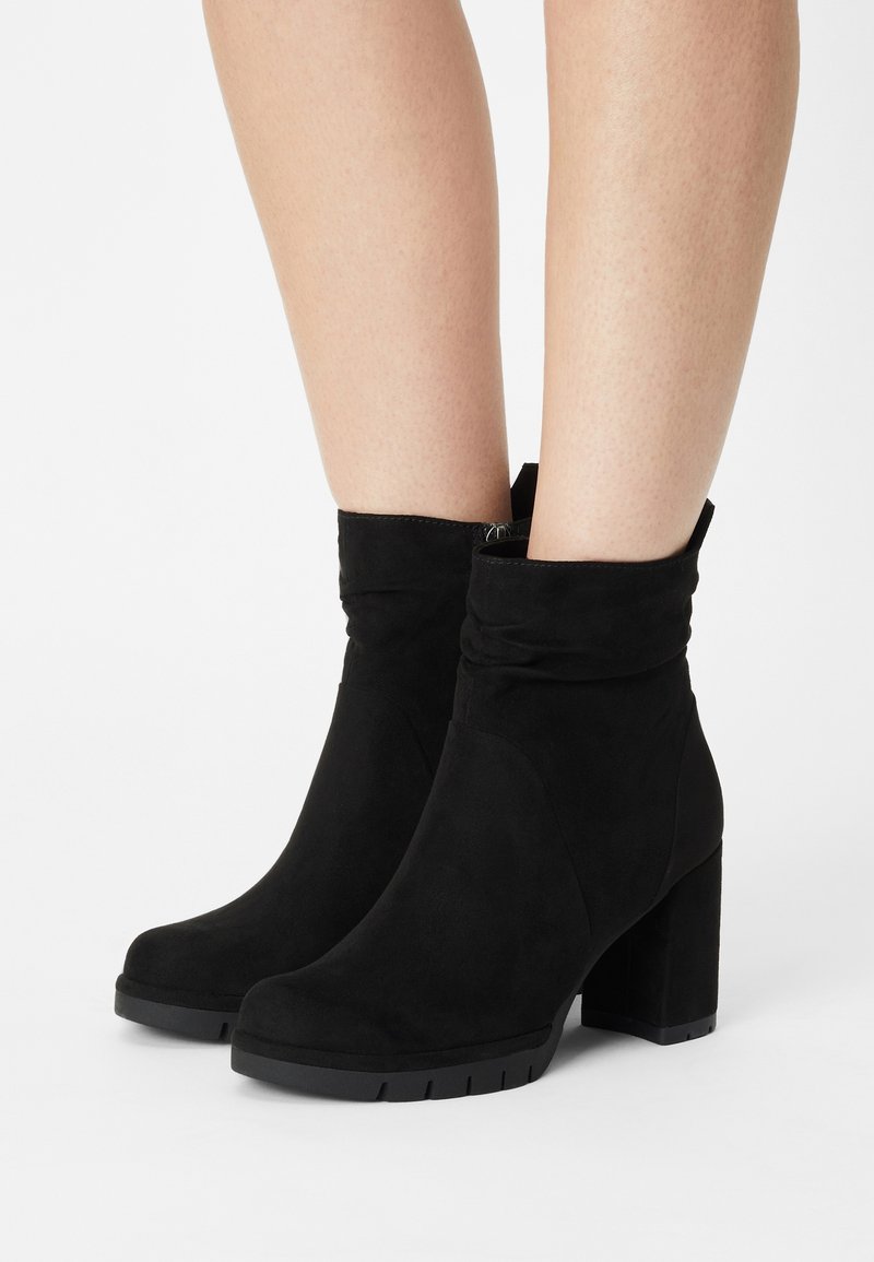 Bottines en suede noir avec un design plié, un talon épais haut et une semelle en caoutchouc. Texture douce et style minimaliste.