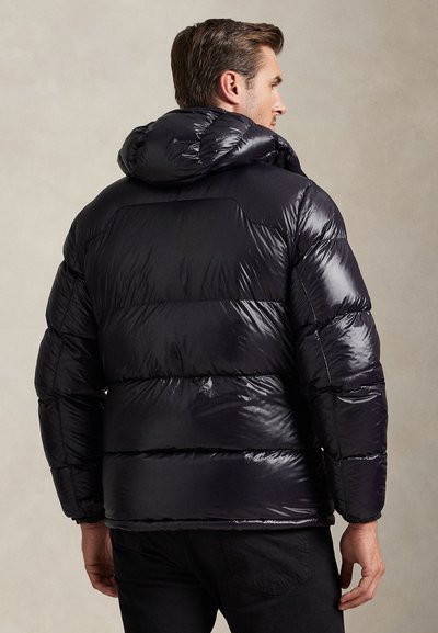 Man wearing a shiny black puffer jacket with a hood, viewed from behind against a plain beige background.