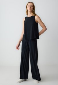Donna in piedi contro uno sfondo neutro, che indossa un top senza maniche nero, pantaloni ampi neri e sneakers bianche.