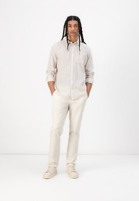 Camisa de botones color beige claro con una textura sutil, combinada con pantalones color crema y zapatillas beige, de pie sobre un fondo blanco.