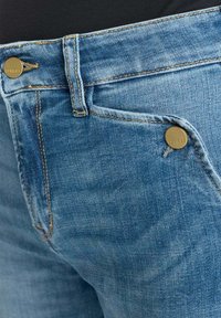 Jeans en denim bleu clair avec une texture délavée, présentant des détails en doré et des coutures discrètes. Comprend des poches avant et des accents de boutons.