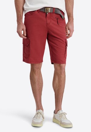 Mann trägt rote Cargoshorts mit mehreren Taschen, einen grauen Gürtel, weißes T-Shirt und weiße Sneakers, steht vor einem einfarbigen Hintergrund.