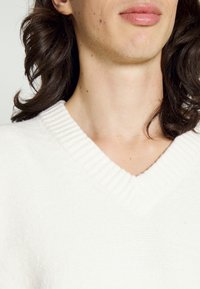 Bílý svetr s výstřihem do V a žebrovaným límcem, vyrobený z měkké látky. Má texturovaný pletený vzor a volný střih.