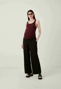 Grūtniecības topiņš bordo krāsā, apvienots ar melnām platiem biksēm. Modelis valkā arī melnas platformas sandales, izceļot minimālisma stilu.