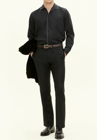 Maglia nera a maniche lunghe con piping bianco a contrasto, abbinata a pantaloni neri sartoriali e una cintura in pelle marrone. Una giacca nera è appoggiata su un braccio.