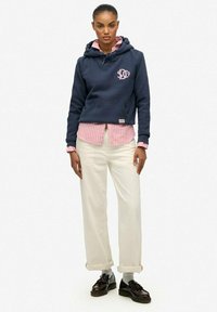 Sudadera con capucha azul marino con logo rosa, llevada sobre una camisa de rayas rosa claro y blanco. Combinada con pantalones blancos y zapatos negros. Fondo blanco.