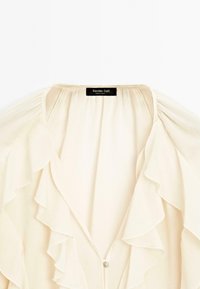 Blusa crema con volant sovrapposti sul davanti, caratterizzata da una morbida texture del tessuto e una piccola chiusura a bottone sul collo. Etichetta del marchio visibile.