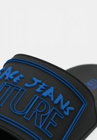 Sort slip-on sandal med en opphøyd blå logo-patch. Mykt gummimateriale med jevn tekstur. Blå detaljer langs kantene på stroppen.