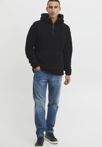 Svart fleece hettegenser med teksturert overflate, halvglidelås, snøring på hetta og frontlomme, kombinert med lyseblå denimbukser og svarte joggesko.