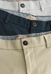 Drei Paar Hosen: hellgrau, marineblau und beige. Aus Baumwollstoff, lockere Passform. Jede hat einen eigenen Knopf und ein Etikett.