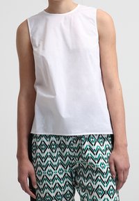 Persona che indossa una canotta bianca senza maniche abbinata a pantaloni con motivo a diamante verde e nero su sfondo bianco.
