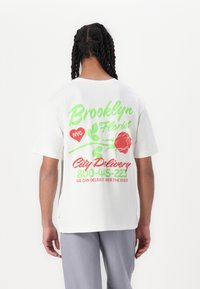 Fehér pamut póló, hátoldalán színes szöveggel és grafikákkal: "Brooklyn Virágárus", egy rózsa és szállítási információ zöld és piros színben.