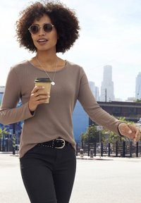 Hellbraunes, geripptes Langarmoberteil, schwarze Skinny-Jeans, silberner Gürtel mit Nieten, runde Sonnenbrille, hält eine Kaffeetasse, städtische Umgebung im Freien.