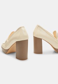 Scarpe con zeppa in pelle verniciata beige, dalla punta arrotondata e con un chunky tacco in legno a righe. Superficie liscia con linee pulite e accenti minimi.