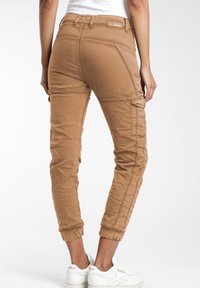 Los pantalones jogger color caqui presentan un diseño ajustado, puños elásticos, múltiples bolsillos y una tela texturizada, combinados con zapatillas blancas.