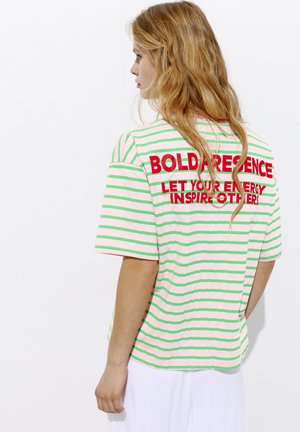 Junge Frau mit langen blonden Haaren, die ein pink-grün gestreiftes Shirt mit rotem Schriftzug "BOLD PRESENCE" und weiße Hose trägt, die nach links schaut.