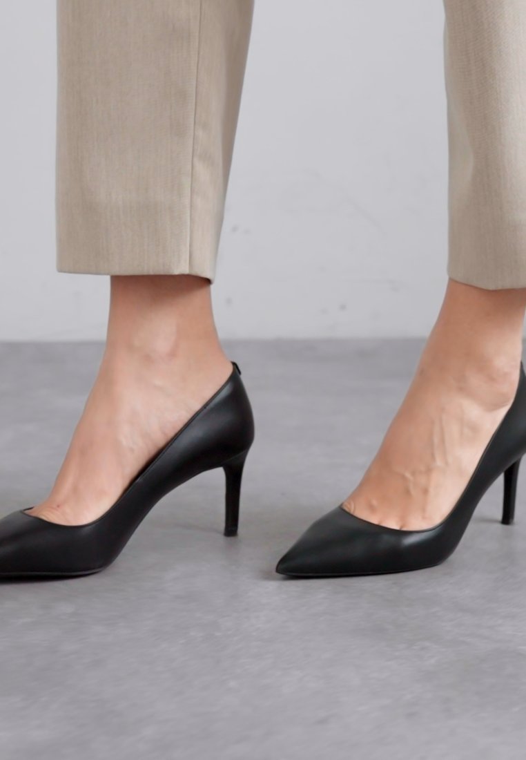 Chaussures à talons hauts en cuir noir avec un design à bout pointu et une silhouette élégante, sur un fond gris clair avec un pantalon aux tons neutres.