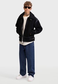 Veste en polaire noire avec un large col, fermeture éclair à l'avant et poches latérales, associée à un jean en denim bleu ample et des baskets blanches.