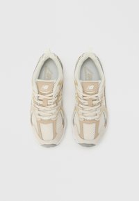 Egy pár bézs és fehér New Balance futócipő hálós és velúr részletekkel, felülnézetből, fehér háttéren.