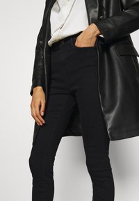 Vaqueros skinny negros con cintura alta, hechos de denim. Combinados con una chaqueta de cuero negra y una blusa blanca, mostrando una silueta elegante.